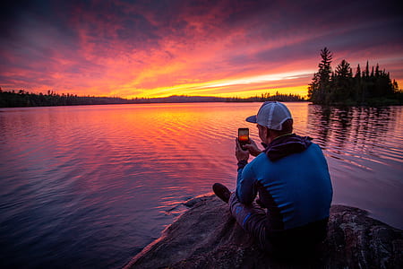man sitting on rock formation front of calm water during golden hour thumbnail