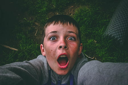 Opened Mouth Black Haired Boy in Gray Full-zip Jacket Standing on Grass Field Taking Selfie thumbnail