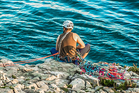 man in brown tank top , black pants and white half face helmet sitting on gray rock formation near body of water thumbnail