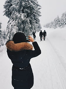 Woman in Black Coat Taking a Picture of Three Person in Front of Her While Walking Through Snow Field thumbnail