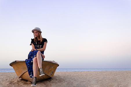 woman in black crew-neck T-shirt and blue skirt sitting on brown boat at the seashore under gray skies during day time thumbnail