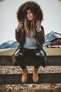 Woman Wearing Brown and Green Parka Sitting on Gray Wooden Fence at Daytime thumbnail
