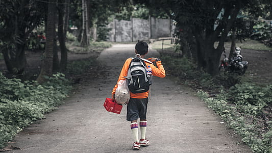 boy in orange long-sleeved shirt carrying backpack walking on pathway thumbnail