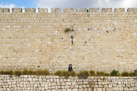two people walking near beige stone wall under gray clouds during daytime thumbnail