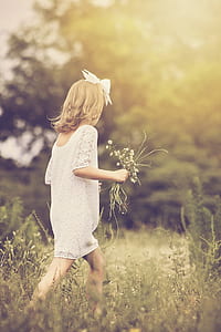 girl wearing white dress holding white flower bouquet while walking on grass field thumbnail