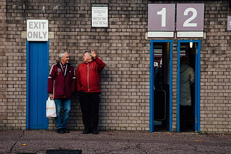 two men leaning on grey brick wall while talking thumbnail