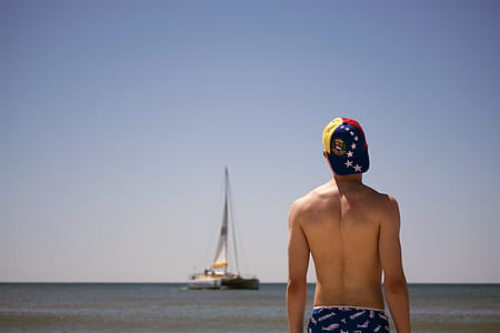 man with yellow, blue, and red cap standing viewing sail boat thumbnail