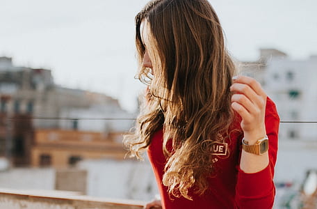 woman wearing red long-sleeved shirt and gold-colored watch with link band stands beside brown concrete fence during daytime thumbnail