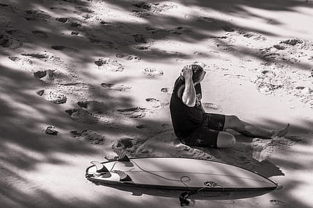 Grayscale Photo of Man Sits Beside Surfboard on Sand thumbnail