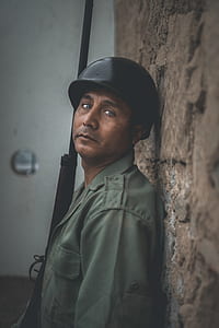 man wearing grey jacket and black helmet holding black rifle leaning on brown concrete wall thumbnail