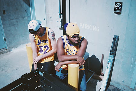 two men wearing Los Angeles Lakers and Golden State Warriors jersey shirts sitting in front of yellow bollards at daytime thumbnail