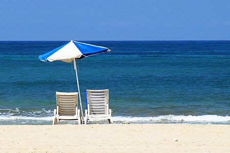 two white chairs under blue and white umbrella near sea under blue clouds during daytime thumbnail