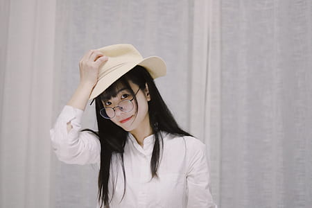 black haired woman wearing clear eyeglasses and white long-sleeved shirt holding brown hat thumbnail
