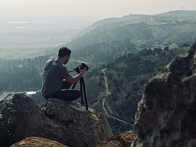 man sitting on gray rock holding black DSLR camera with tripod thumbnail