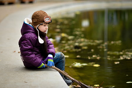 boy sitting on pavement while holding stick in front of body of water thumbnail