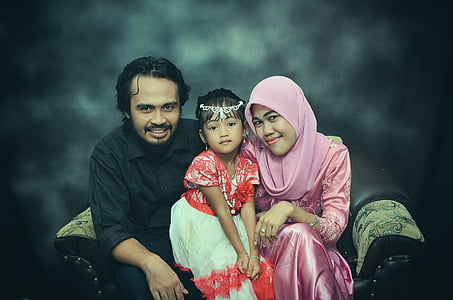 man sitting beside girl wearing red dress and woman wearing pink hijab thumbnail
