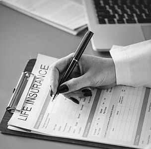 grayscale photo of person's right hand holding pen and about to write on Life Insurance paper beside laptop computer thumbnail