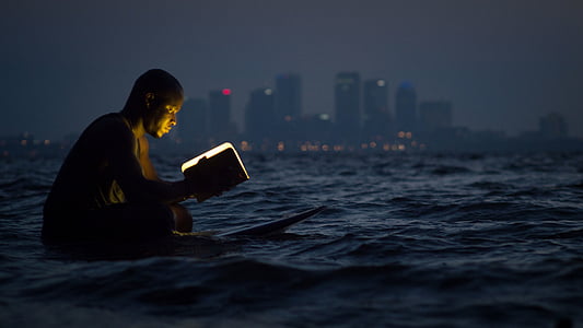 man reading book while sitting on surfboard thumbnail