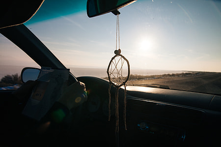 dreamcatcher hanged from rear view mirror inside car driving on road along shore during golden hour thumbnail
