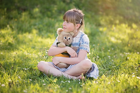 girl hugging brown bear plush toy while sitting on grass field during daytime thumbnail