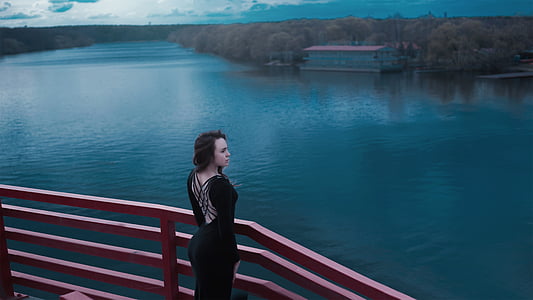 woman in black cage-back dress standing near wooden railings surrounded with body of water thumbnail