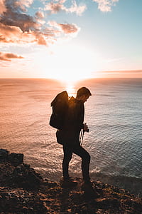 man wearing black backpack standing on cliff beside ocean during orange sunset thumbnail