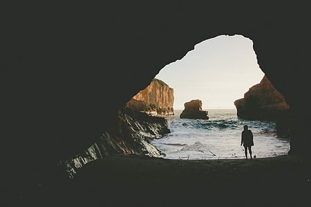silhouette of person standing under cave near seashore during daytime thumbnail
