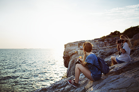 woman taking photo of woman while sitting on cliff near body of water during daytime thumbnail