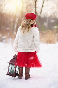 girl in white cardigan and red dress holding lantern standing on snow field during daytime thumbnail