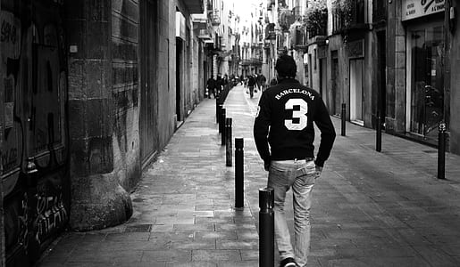 Grayscale Photo of Man Wearing Long-sleeved Shirt and Pants Walking on Concrete Pathway Near Building thumbnail