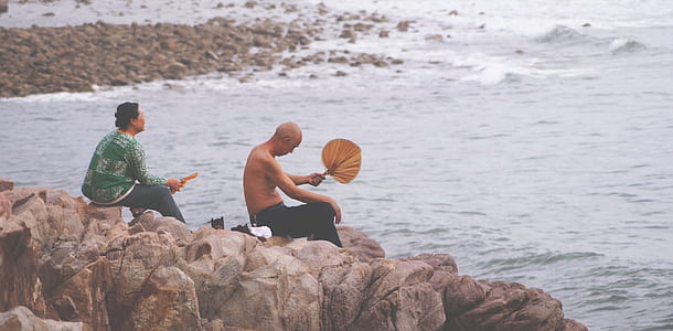 two men sitting on rock beside body of water thumbnail