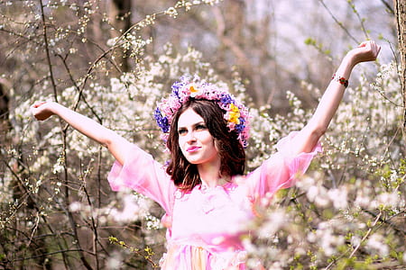 fashion photography of woman wearing pink elbow-sleeved top and multicolored floral headpiece raising both arms surrounded by flowering plants thumbnail