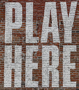 Large painted letters say PLAY HERE in white on an old red brick wall. thumbnail