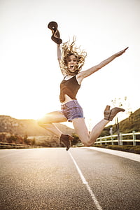 woman jumping on asphalt road while holding bucket hat during daytime thumbnail