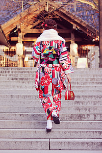 woman in red, white, and black floral long-sleeve traditional dress on stairs facing the brown wooden house thumbnail
