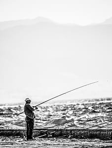 Man Standing Near Seashore Holding Fishing Rod on Grayscale Photography thumbnail